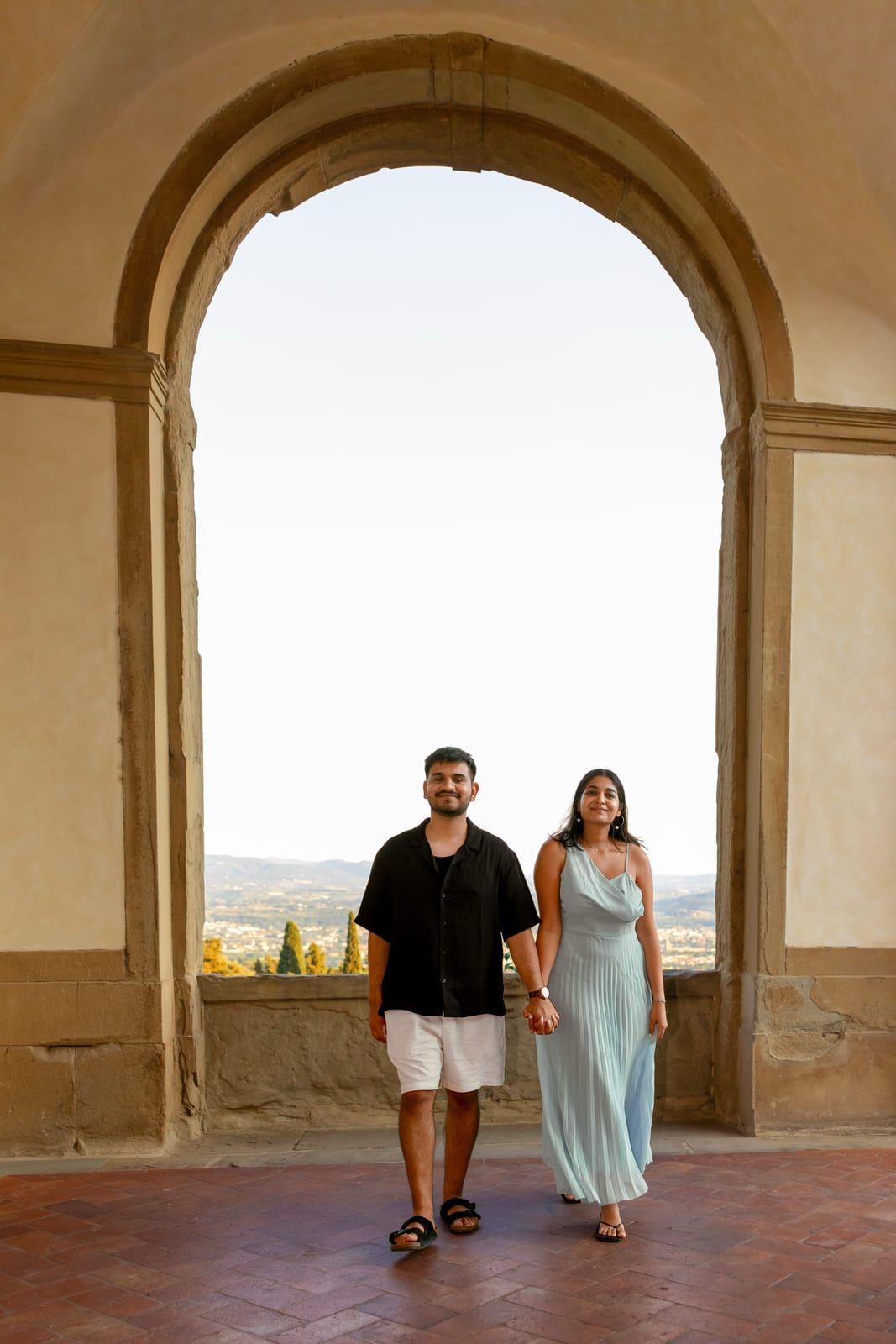Pooja and Barun holding hands beneath a stone arch overlooking the Tuscan hills